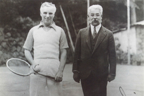 A black-and-white photograph of Charles Chaplin holding a tennis racket, standing next to Shozo Yamaguchi in a suit, on a tennis court